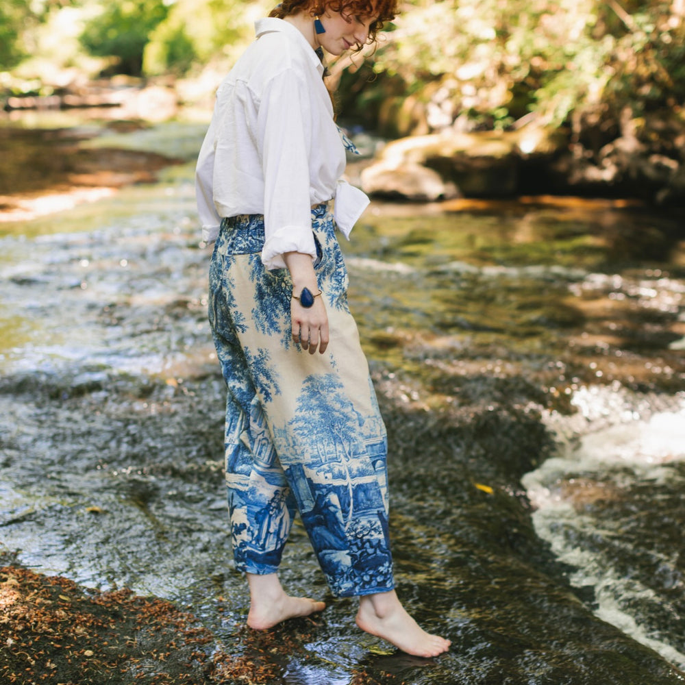 
                  
                    Model wearing Let the Light In linen Artist Pant featuring print of Delft blue and white landscape pottery, and positive message of light and hope - Market of Stars
                  
                