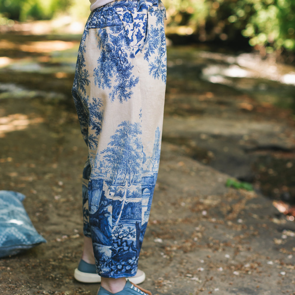
                  
                    Model wearing Let the Light In linen Artist Pant featuring print of Delft blue and white landscape pottery, and positive message of light and hope - Market of Stars
                  
                