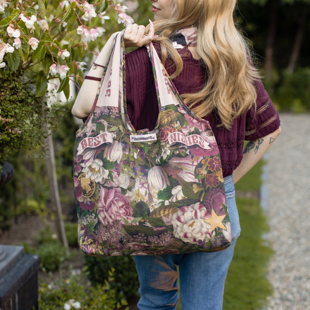 
                  
                    Model holding linen blend market tote featuring Victorian wishbone–vintage coin–evening star–dandelion–and “Best Wishes” banner on floral background – Market of Stars
                  
                