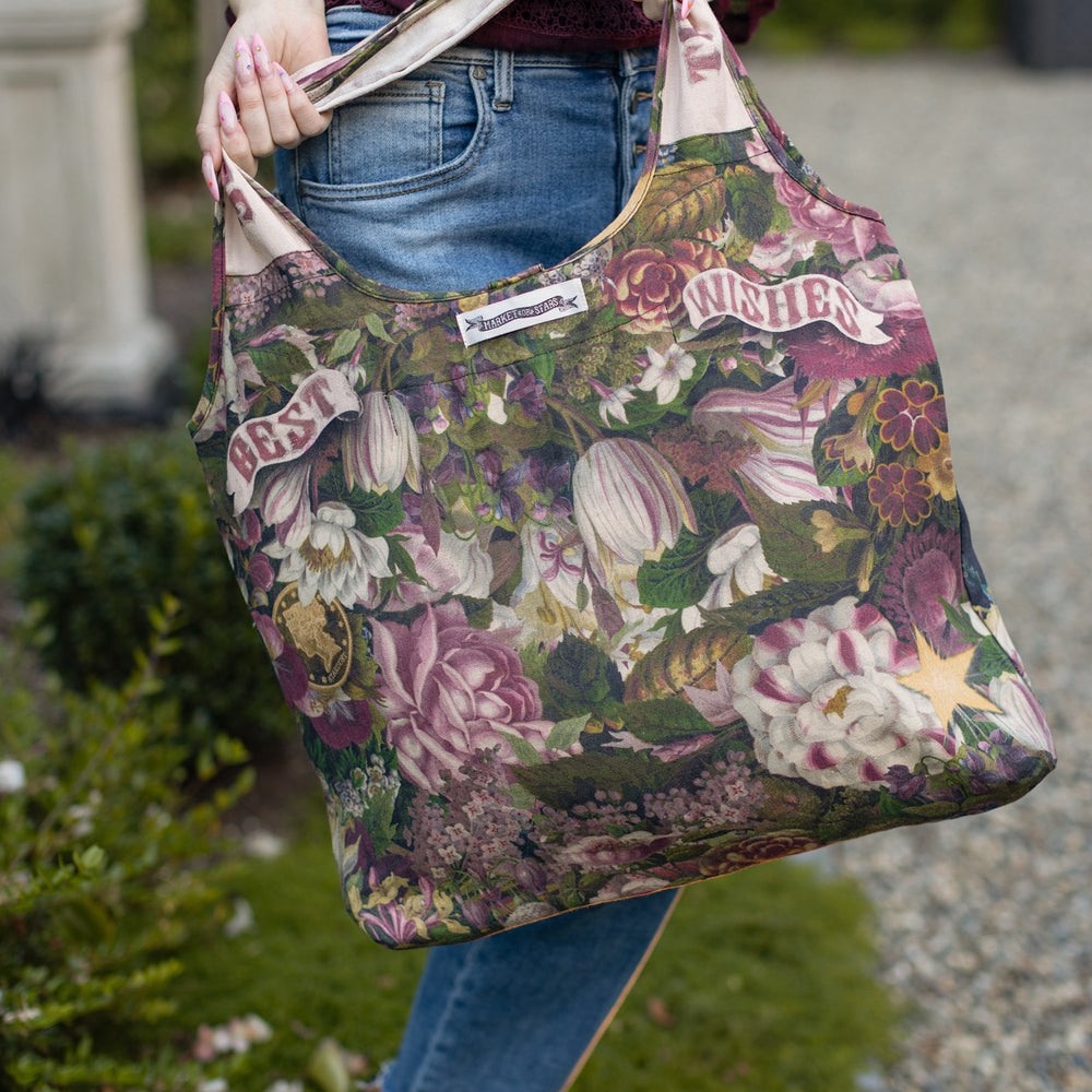 
                  
                    Model holding linen blend market tote featuring Victorian wishbone–vintage coin–evening star–dandelion–and “Best Wishes” banner on floral background – Market of Stars
                  
                