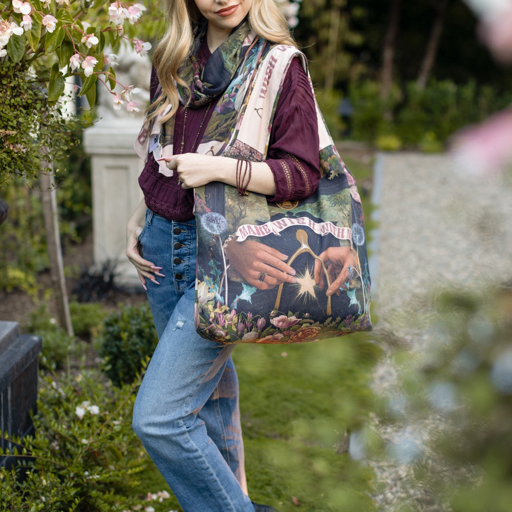 
                  
                    Model holding linen blend market tote featuring Victorian wishbone–vintage coin–evening star–dandelion–and “Best Wishes” banner on floral background – Market of Stars
                  
                