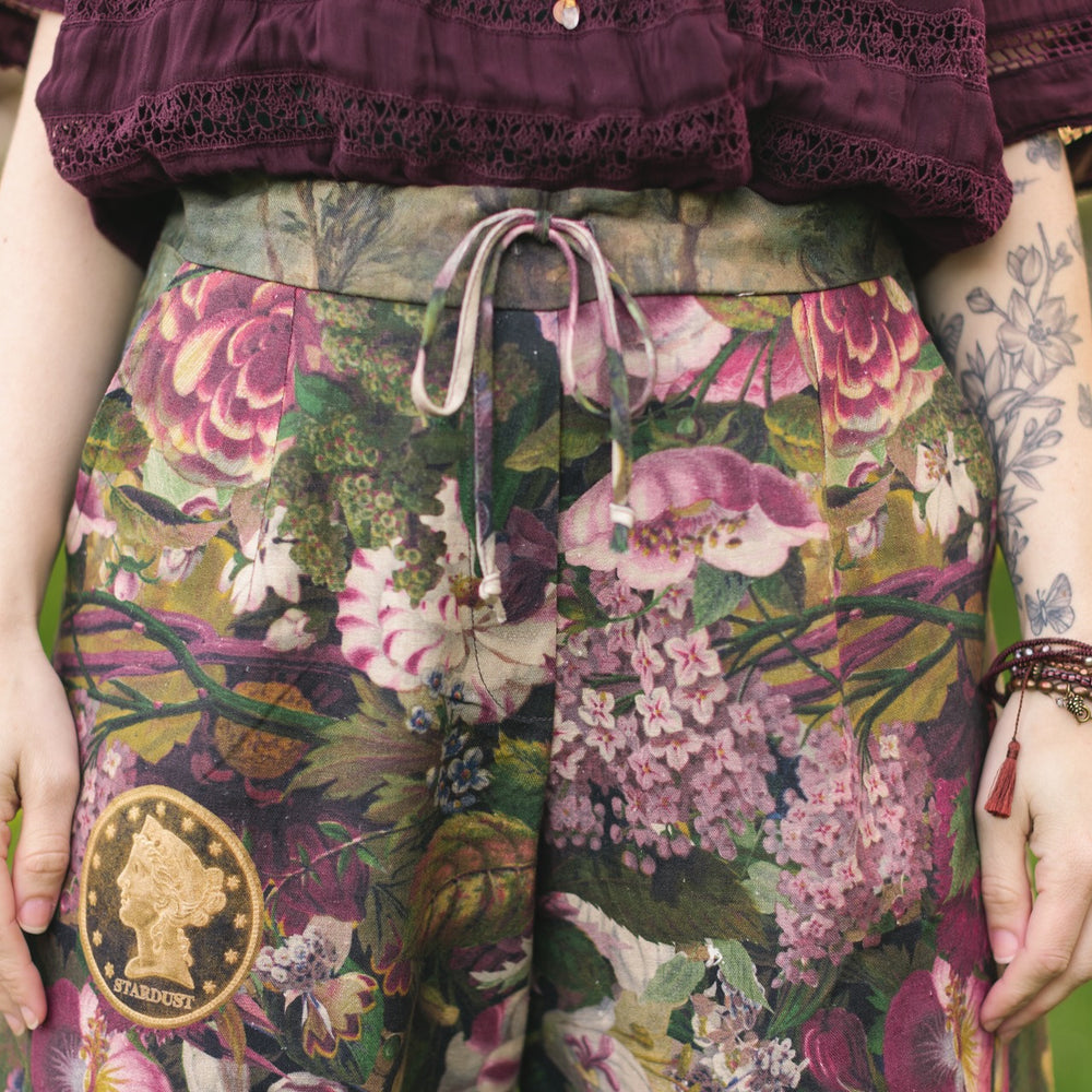 
                  
                    Model wearing linen blend artist pants featuring antique wishbone–Victorian coin–evening star–dandelion art and “Wish with Me” banner on floral background – Market of Stars
                  
                