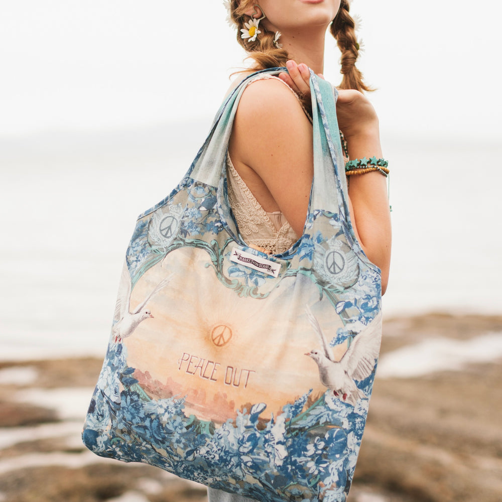 Model wearing our Peace Out tote bag featuring 1800's perfumeur sign with gentle tones of blue & aqua, landscapes, clouds, dove, and peace symbols -Market of Stars.