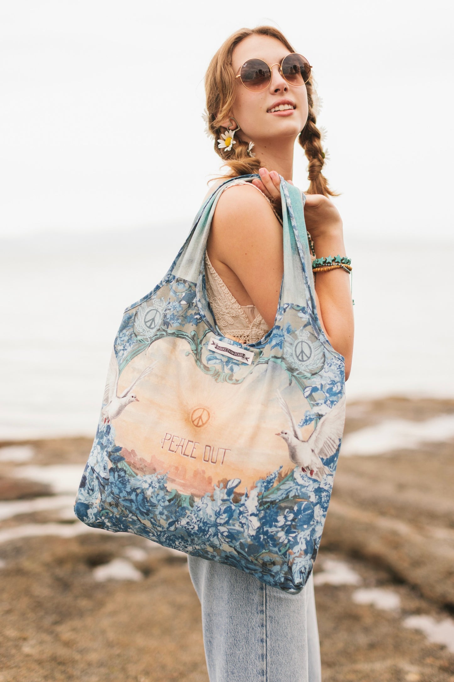 Model wearing our Peace Out tote bag featuring 1800's perfumeur sign with gentle tones of blue & aqua, landscapes, clouds, dove, and peace symbols -Market of Stars.