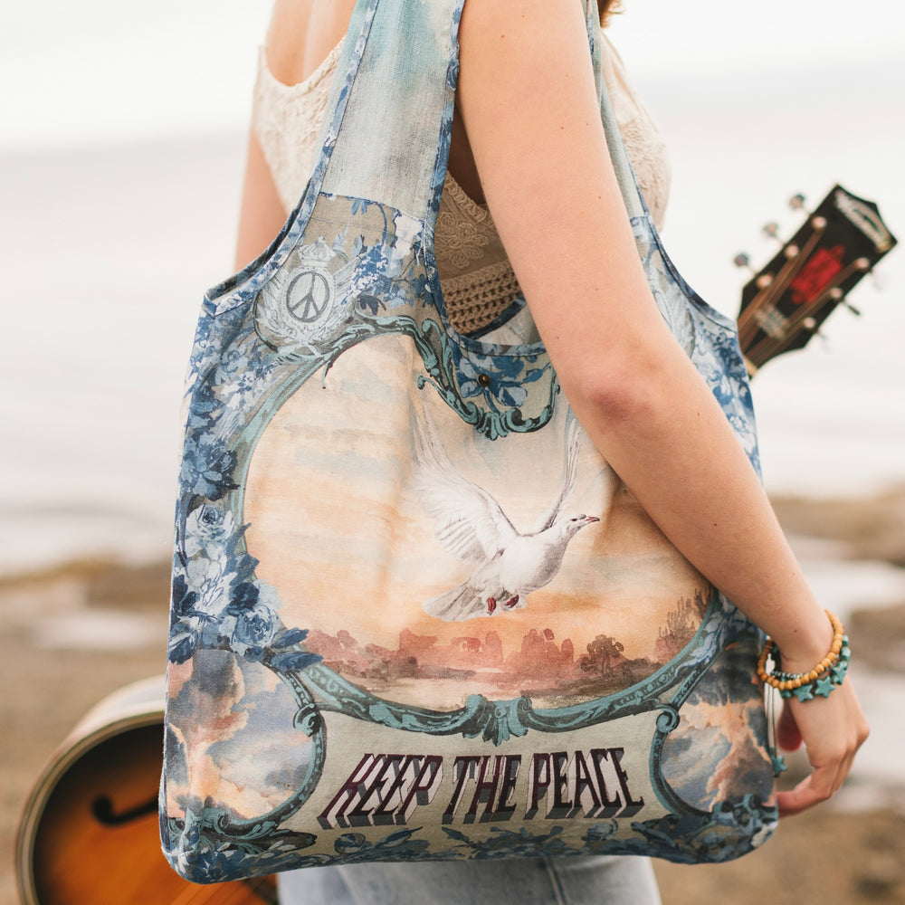 
                  
                    Model wearing our Peace Out tote bag featuring 1800's perfumeur sign with gentle tones of blue & aqua, landscapes, clouds, dove, and peace symbols -Market of Stars.
                  
                