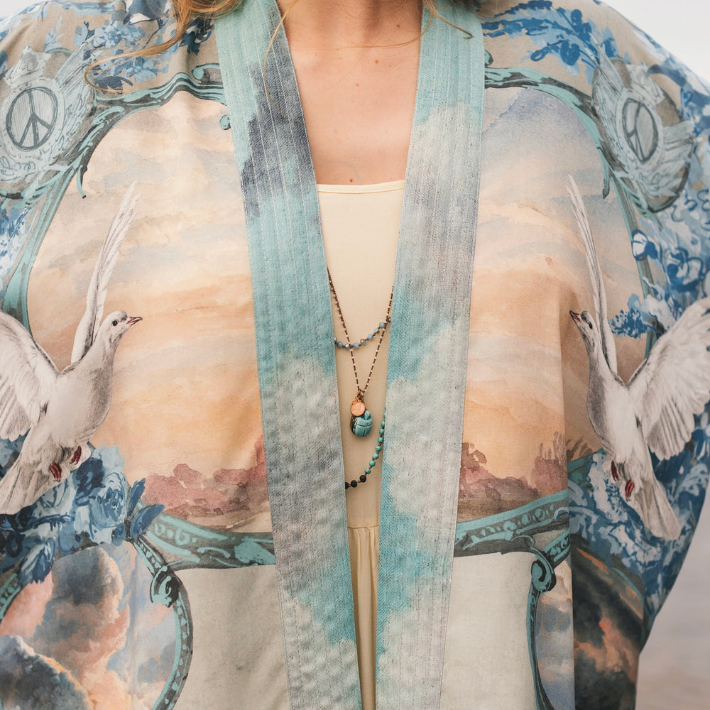 
                  
                    Model wearing our Peace Out Pixie Duster featuring 1800's perfumeur sign with gentle tones of blue & aqua, landscapes, clouds, dove and peace symbols -Market of Stars.
                  
                