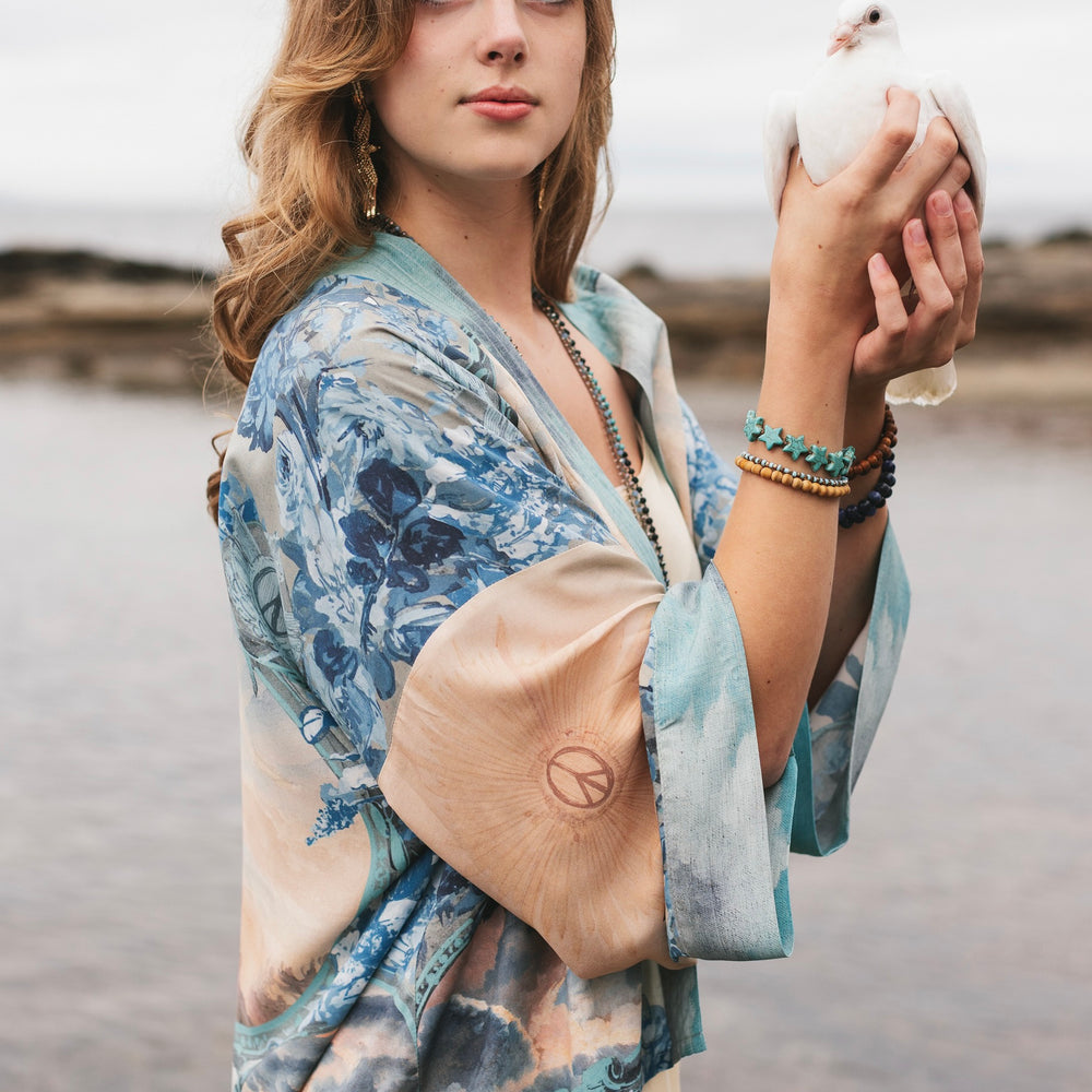 
                  
                    Model wearing our Peace Out Pixie Duster featuring 1800's perfumeur sign with gentle tones of blue & aqua, landscapes, clouds, dove and peace symbols -Market of Stars.
                  
                