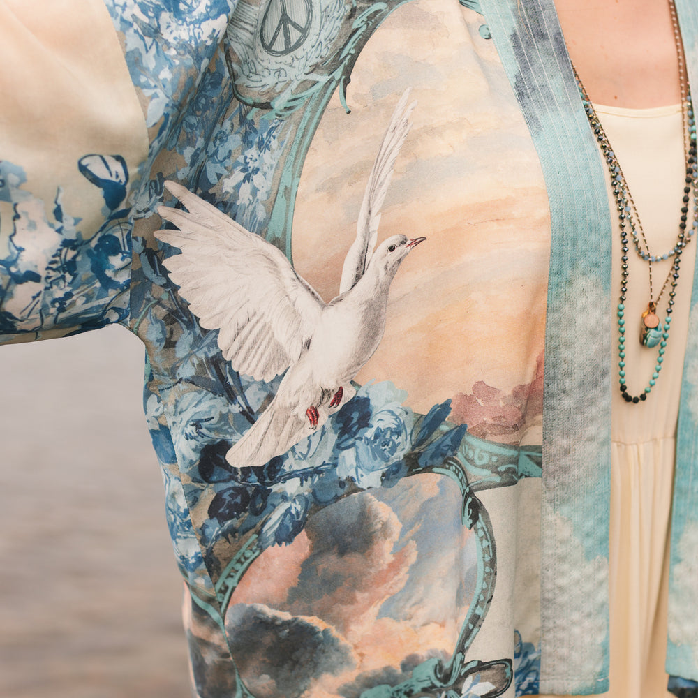 
                  
                    Model wearing our Peace Out Pixie Duster featuring 1800's perfumeur sign with gentle tones of blue & aqua, landscapes, clouds, dove and peace symbols -Market of Stars.
                  
                