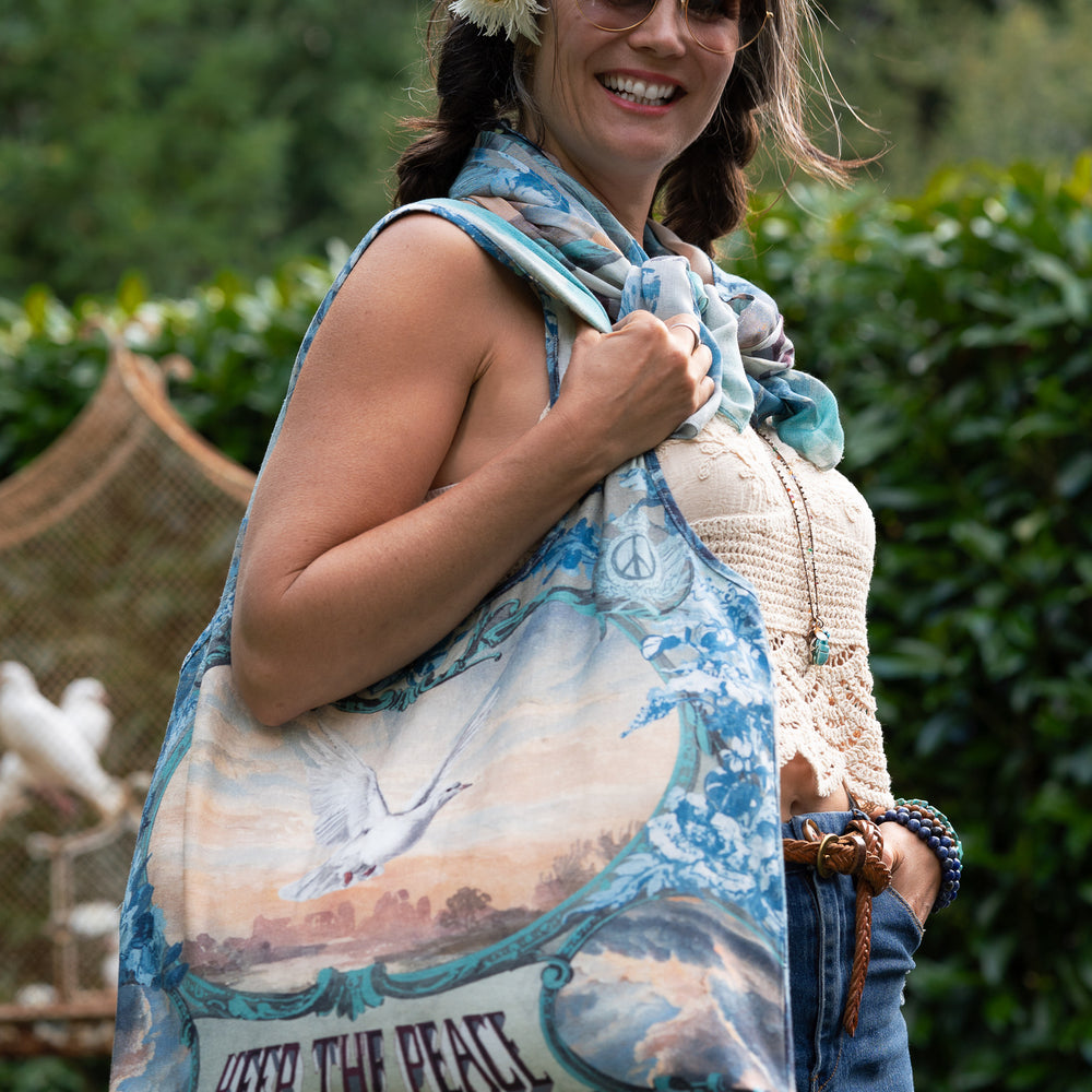 
                  
                    Model wearing our Peace Out tote bag featuring 1800's perfumeur sign with gentle tones of blue & aqua, landscapes, clouds, dove, and peace symbols -Market of Stars.
                  
                