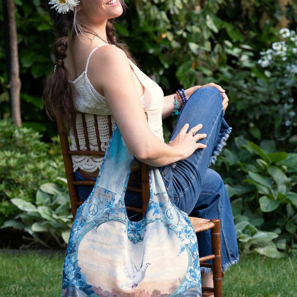 
                  
                    Model wearing our Peace Out tote bag featuring 1800's perfumeur sign with gentle tones of blue & aqua, landscapes, clouds, dove, and peace symbols -Market of Stars.
                  
                