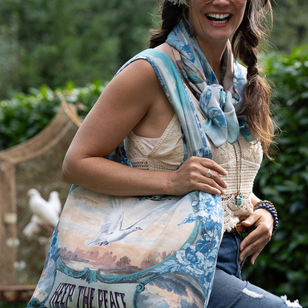 
                  
                    Model wearing our Peace Out tote bag featuring 1800's perfumeur sign with gentle tones of blue & aqua, landscapes, clouds, dove, and peace symbols -Market of Stars.
                  
                
