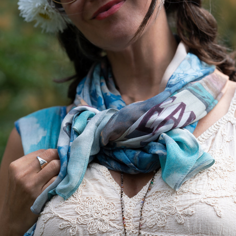 
                  
                    Model wearing our Peace Out scarf featuring 1800's perfumeur sign with gentle tones of blue & aqua, landscapes, clouds, dove, and peace symbols -Market of Stars
                  
                