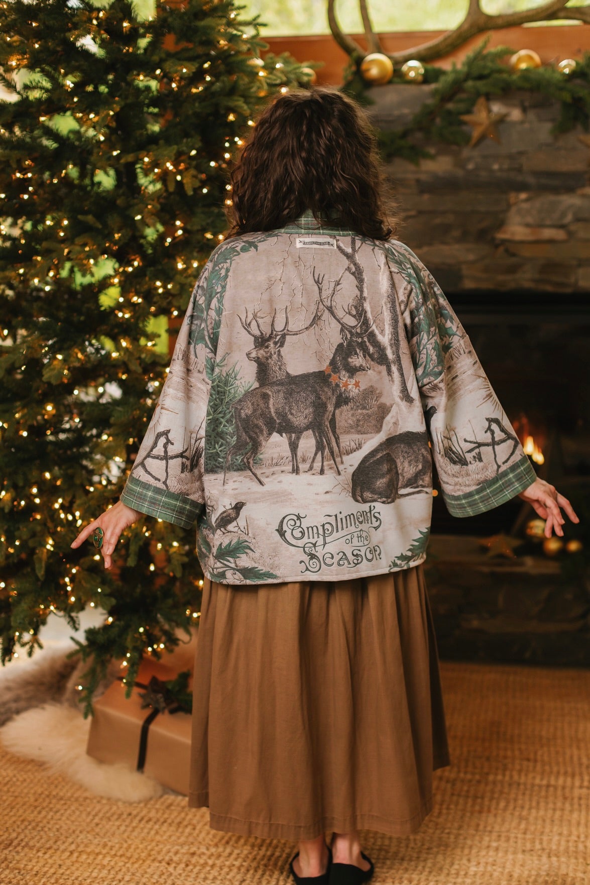 Model wearing a fleece lined jacket with pockets with Victorian-era images from holiday greeting cards — including reindeer in the snow, a little fir tree, golden stars, and traditional plaid trim - Market of Stars